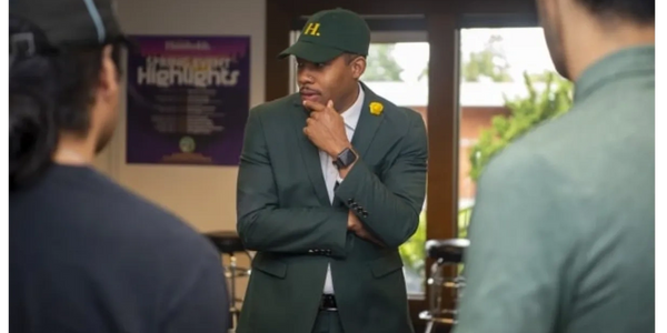 Man in green suit and cap thoughtfully listens to two people indoors.