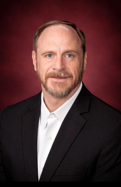 Professional headshot of a man in a black suit and white shirt against a burgundy background.
