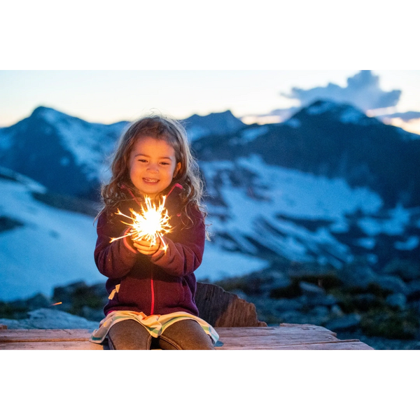 A young girl happily holding a glowing sparkler in a snowy mountain setting at dusk.