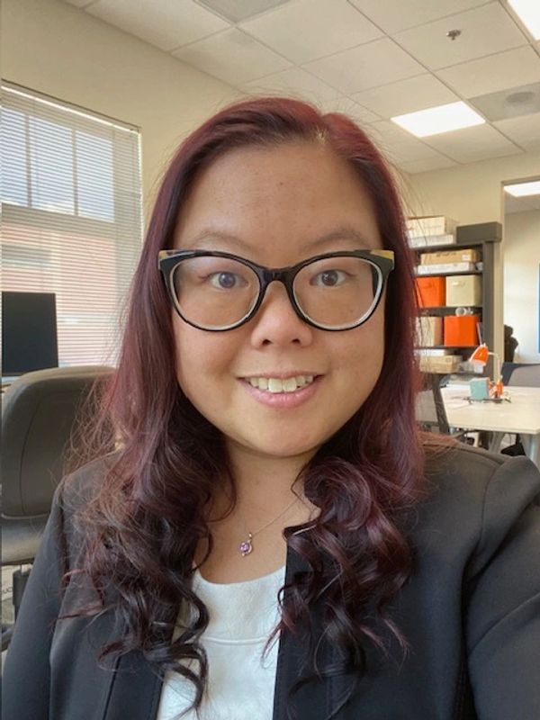 An Asian-American woman who wears glasses and is smiling while wearing a black suit jacket. 