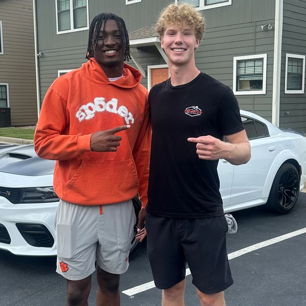 Two smiling men posing outside near a white car.