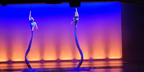 Two aerial silk performers on stage with blue and orange lighting.