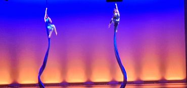 Two aerial silk performers on stage with blue and orange lighting.