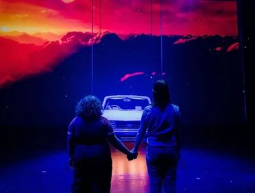 Two people holding hands facing a screen with a car and sunset.
