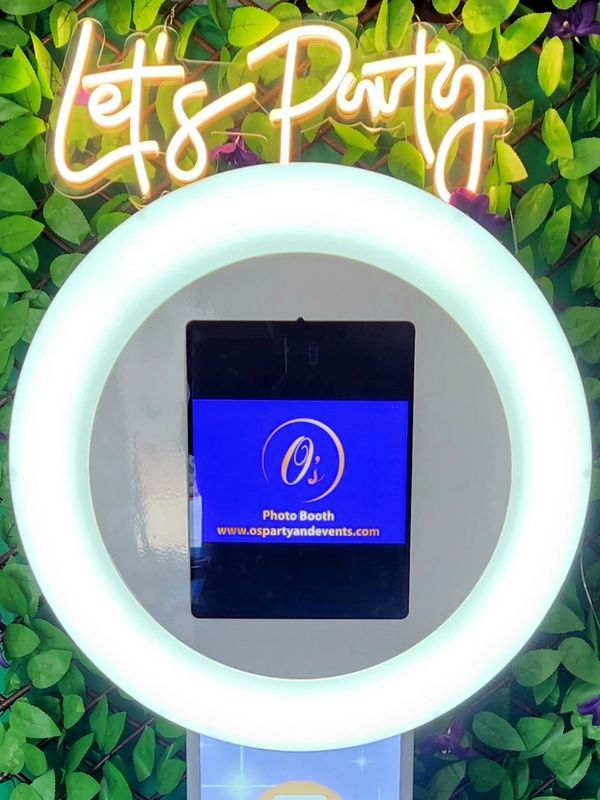Photo booth with glowing ring light and "Let's Party" neon sign on leafy backdrop.
