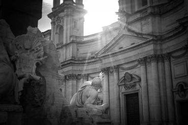 Black and white photo of a statue reaching towards sunlight near a classical building.