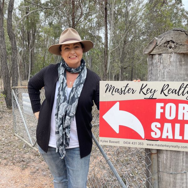 Woman smiling next to a real estate 'For Sale' sign in a wooded area.