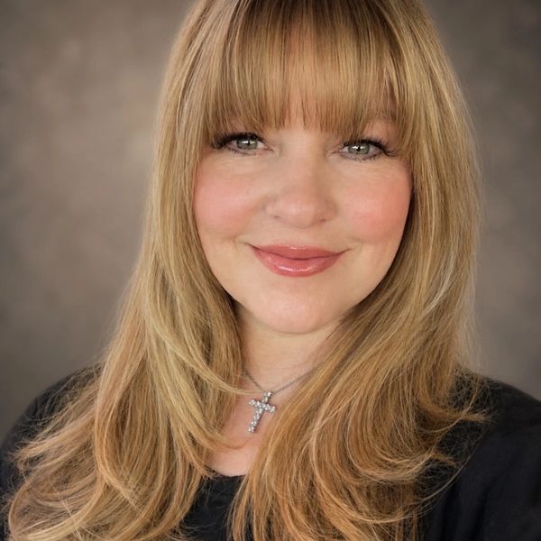 Smiling woman with blonde hair and bangs wearing a cross necklace.