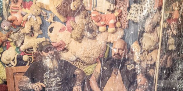 Two tattooed men sitting on a couch with a wall full of stuffed animals behind them.