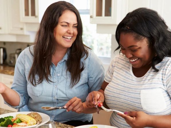 Two women laughing and sharing food in a kitchen.