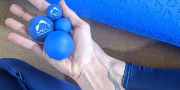 Hand holding four blue MELT therapy balls with a blue foam roller in the background.