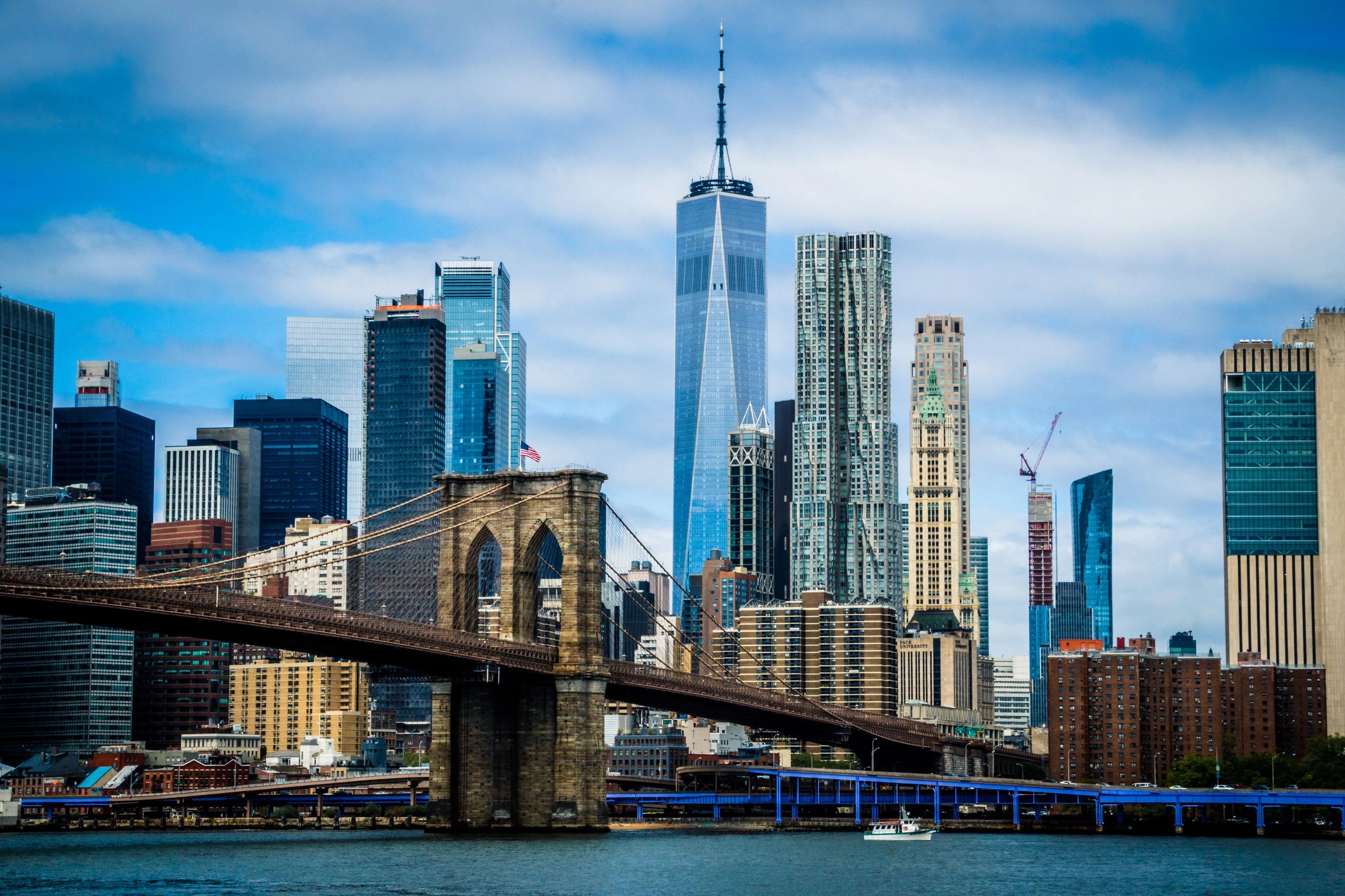 Brooklyn Bridge and Hudson River in front of New York City skyline and cloudy blue sky