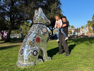 A man holding a child near a mosaic dog sculpture in a park.