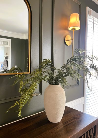 White vase with green ferns on a wooden table against a dark green wall.
