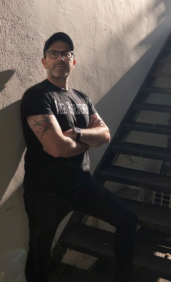 Man in glasses and cap leaning against a wall, arms crossed, standing next to a staircase.