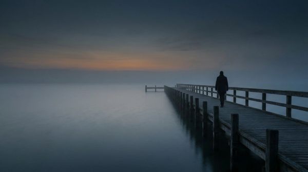 A solitary figure walks on a foggy pier at dusk.