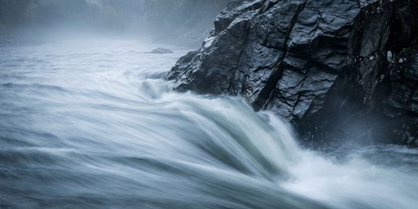 Rushing water flows rapidly past rocky cliffs under a misty sky.
