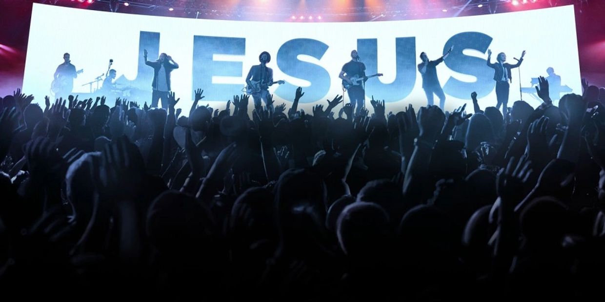Live worship concert with band and crowd raising hands in front of JESUS sign.