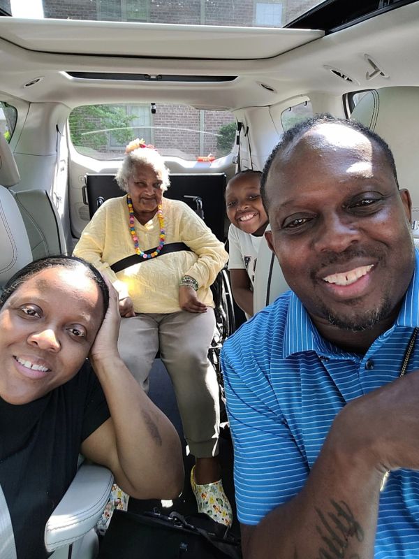 Happy family smiling inside a car with an elderly woman in a wheelchair.