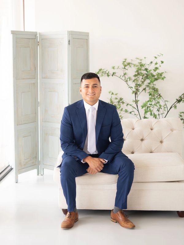 A man in a blue suit sitting on a beige couch in a bright room.