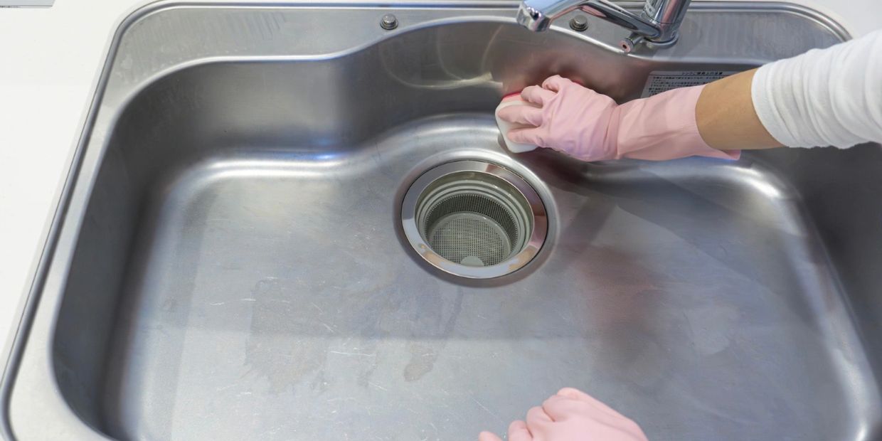 A person wearing pink gloves cleaning a stainless steel sink with a sponge.
