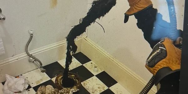 A gloved person pulling out a large, dirty clog from a drain in a checkered floor room.
