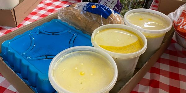 Takeout meal with soup, bread, and a blue container on a checkered tablecloth.