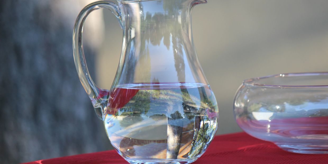 Clear glass pitcher filled with water on a red tablecloth, reflecting outdoor scenery.