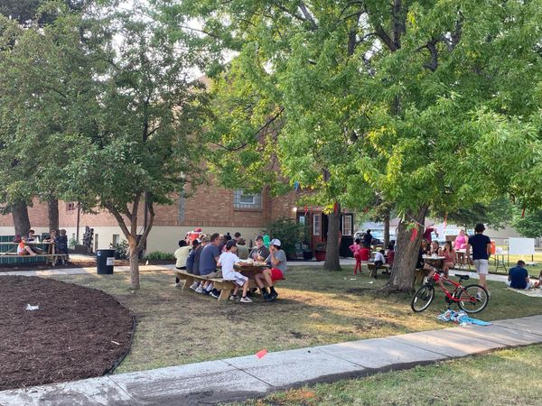 People gathered outdoors at picnic tables under trees near a brick building.