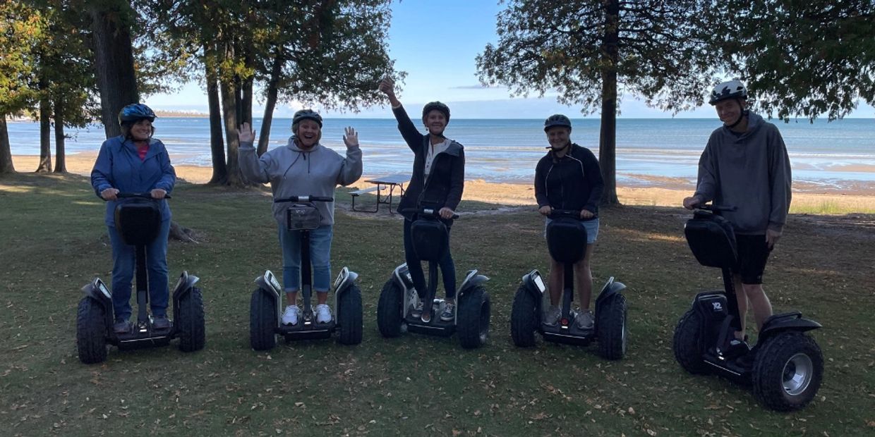 Group of five people riding Segways near a beach with trees and grass.