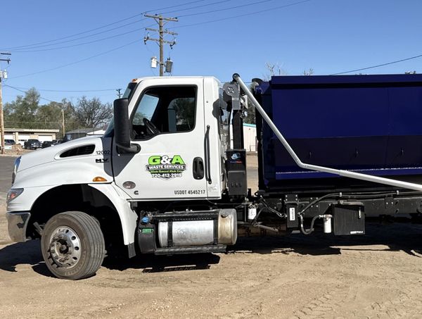 White and blue waste service truck parked on dirt under clear sky.