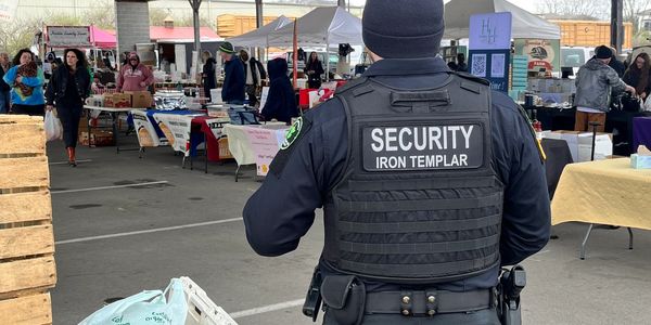 Security guard at a busy outdoor market wearing a vest labeled 'IRON TEMPLAR'.