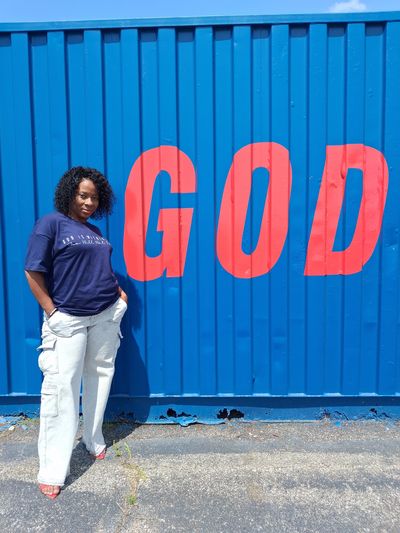 Woman posing by a blue wall with "GOD" in red letters.