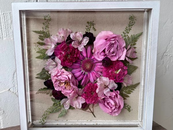 Framed arrangement of pink and purple flowers with green ferns.