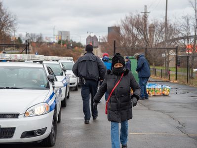 A woman walks past police cars on a cold day with people and supplies in the background.