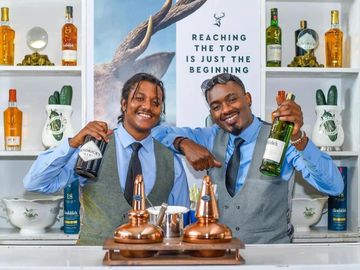 Two bartenders smiling and holding bottles behind a bar with a motivational sign.