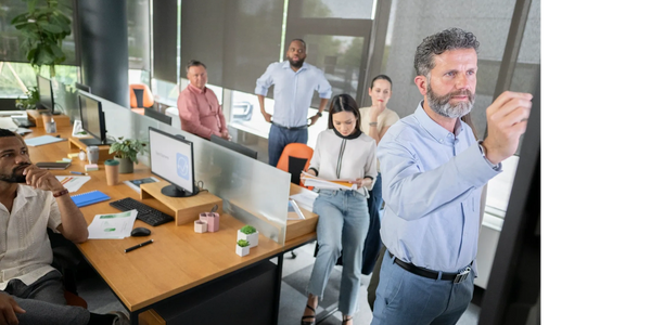 A man leads a meeting, writing on a board while colleagues observe and review documents.