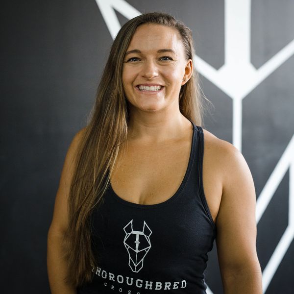 Smiling woman in a black tank top with a gym logo stands against a geometric black and white wall.