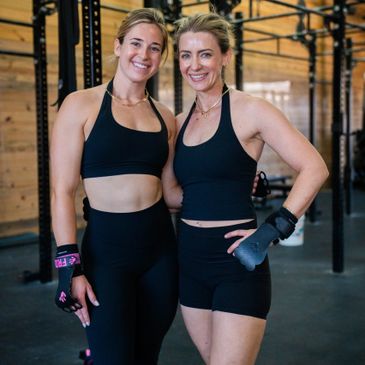 Two women in black workout gear smiling in a gym.