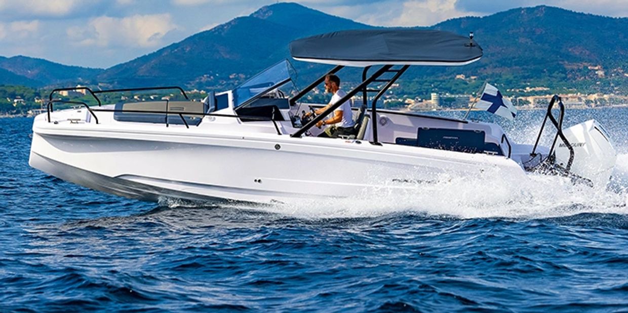A man drives a sleek white motorboat on blue waters with mountains in the background.