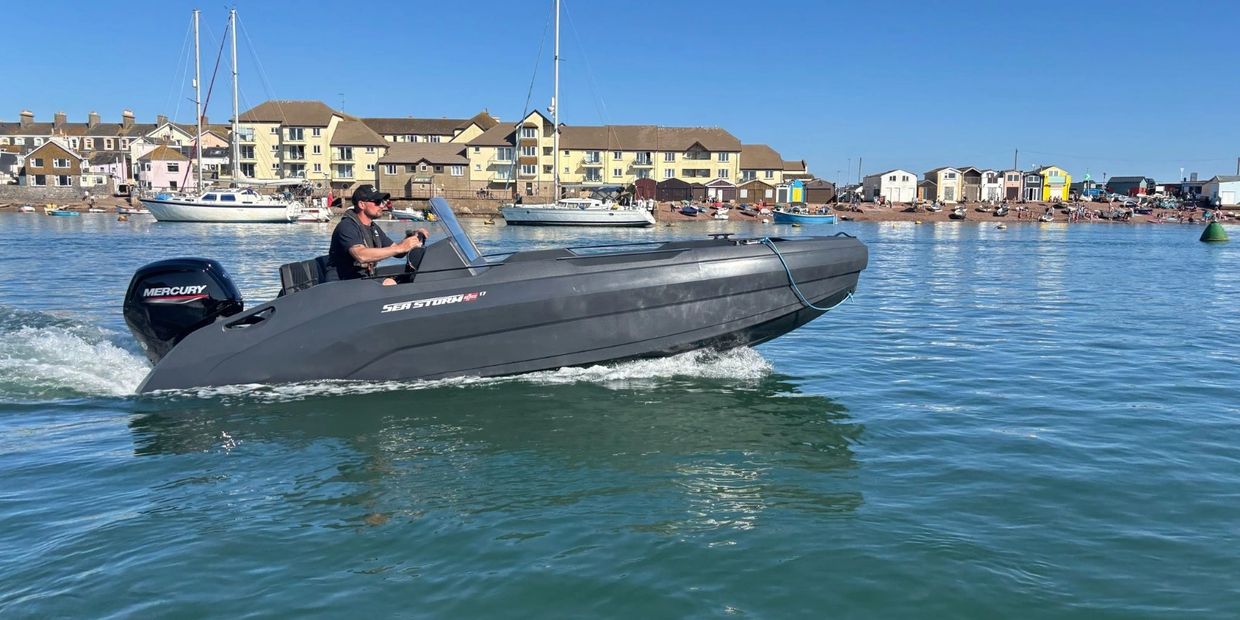 Man steering a sleek black Sea Storm boat on calm water near a coastal town.