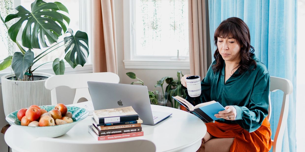 A woman reads a book and drinks coffee at a white table with a laptop and books.