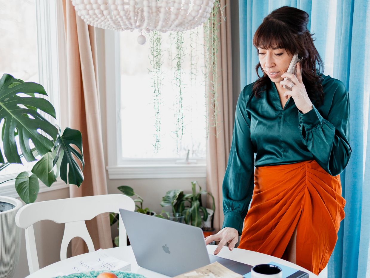 Woman in teal blouse and orange skirt talks on phone while using laptop at home.