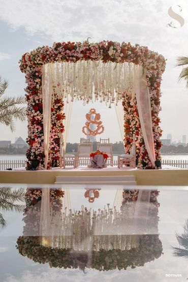Elegant floral wedding mandap with reflection over water under a cloudy sky.