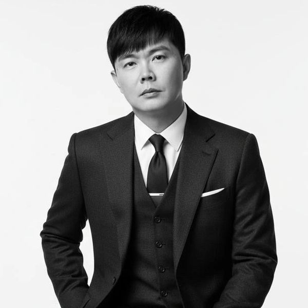 Man in a dark suit sitting on a stool against a plain background.