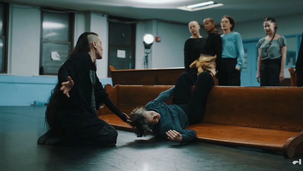 Person with unique hairstyle helps lying person in a group setting.