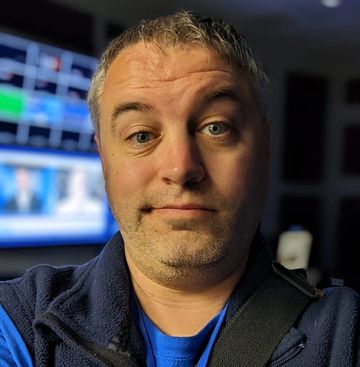 Man taking a selfie in a dimly lit room with screens in the background.