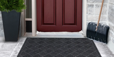 Front door in winter, a contours mat laid in front of the doorway, a shovel propped at the wall.