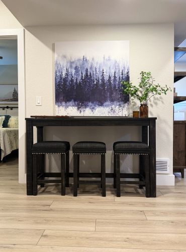 Three black stools under a wooden table with a forest painting above.
