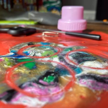 Close-up of colorful paint palette with a brush and a container lid in the background.